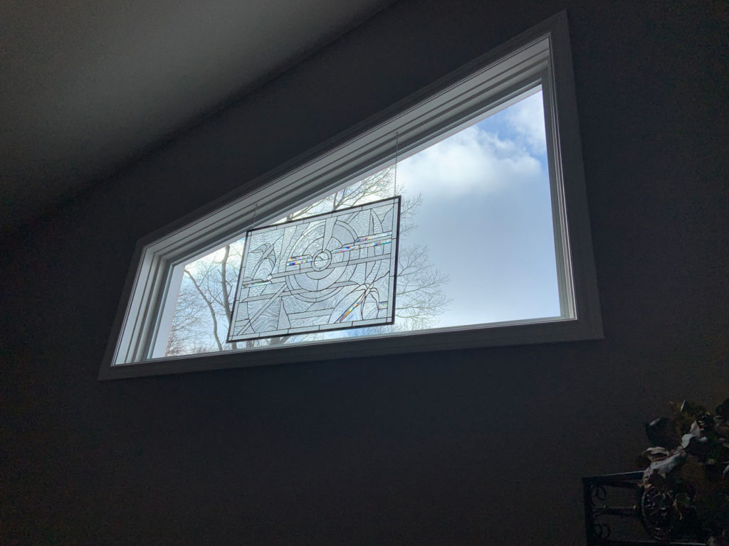 A rectangular stained glass panel featuring a clear, beveled, and textured geometric design with a central circular motif and subtle blue, red, and yellow colored glass accents, suspended by chains within a large, modern triangular window. Outside, a cloudy sky and bare tree branches are visible.