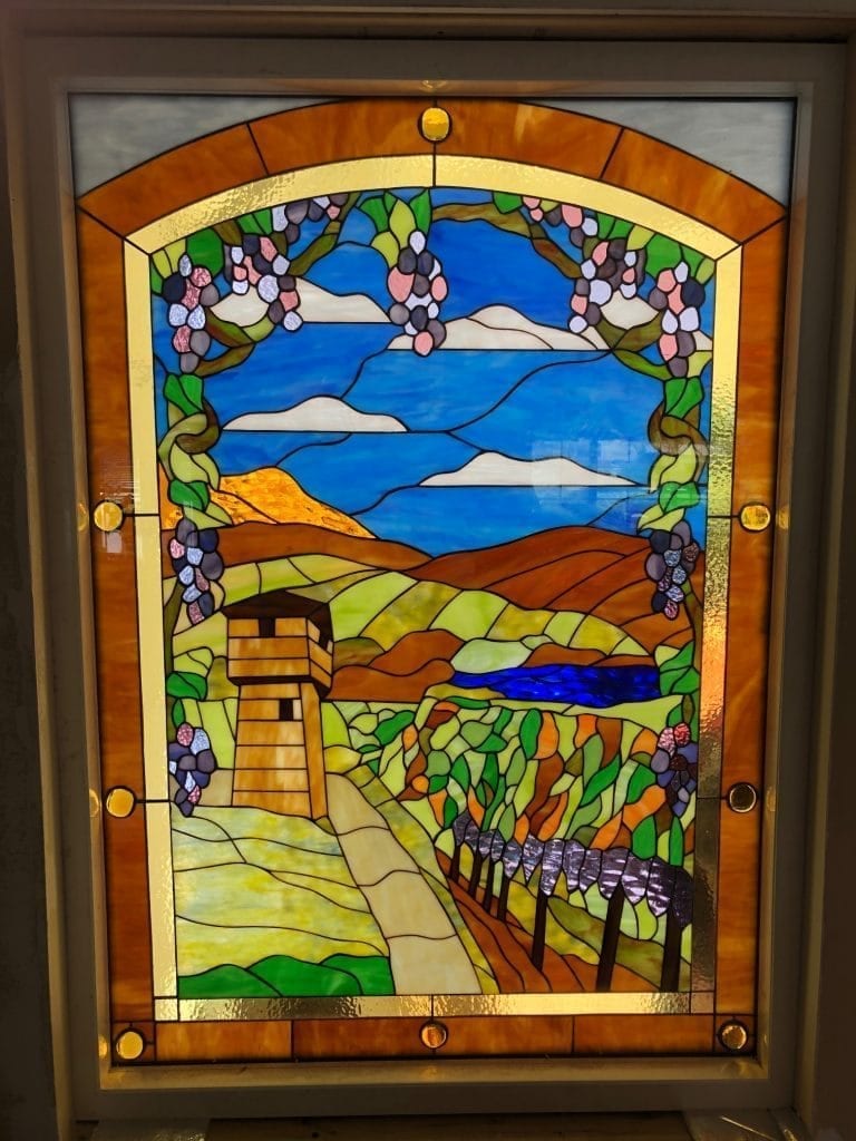 A rectangular, leaded stained glass window panel depicting a vibrant vineyard landscape. The scene includes rolling green and brown hills, a winding path leading to a rustic watchtower, rows of grapevines, a blue river, a blue sky with white clouds, and an arched border with hanging grape clusters and golden glass jewels.