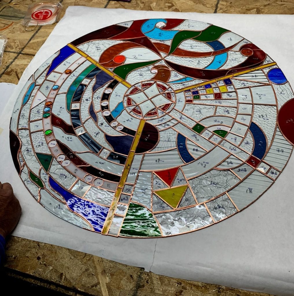 A large, intricate circular stained glass panel lying on a white pattern paper, currently in the copper foiling stage. The design features a central star-like motif surrounded by swirling, abstract geometric patterns made from various types of glass: clear textured, opalescent white, vibrant reds, blues, greens, yellows, ambers, and browns. Several small, round glass jewels are embedded within the clear sections. A human hand is visible in the lower left corner, indicating active construction.