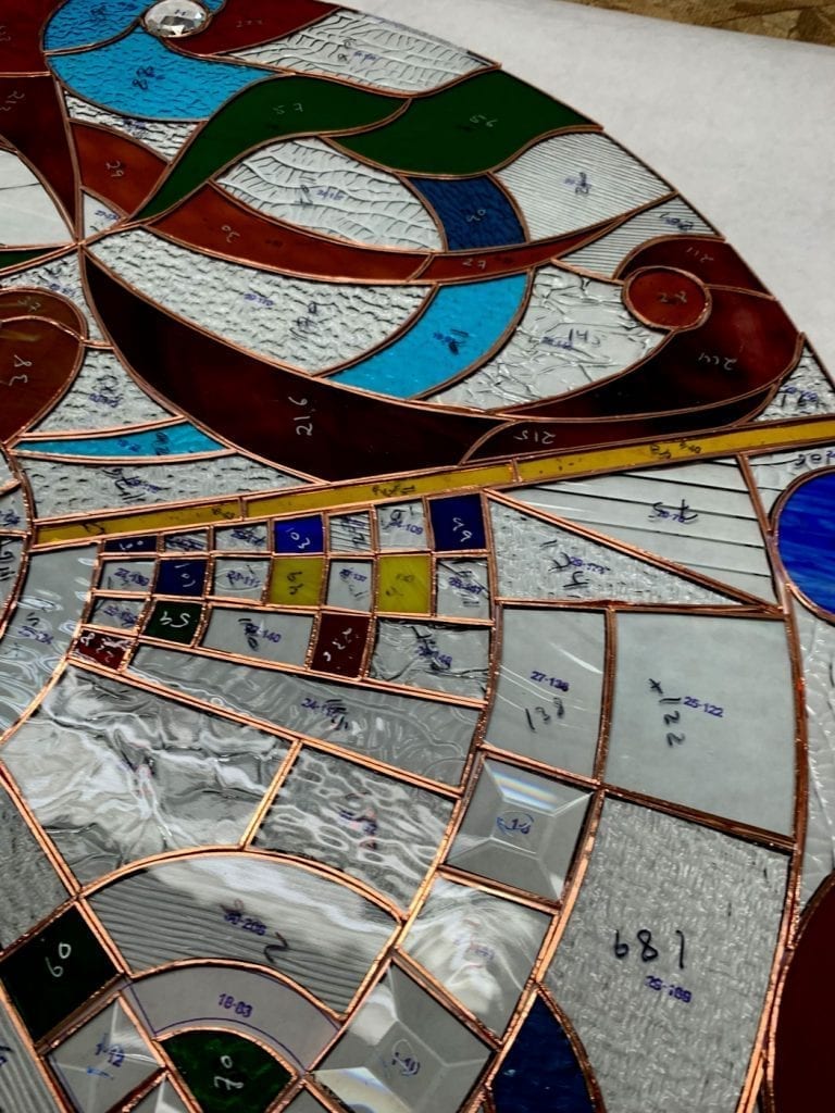 Close-up of an intricate stained glass panel in its assembly stage, utilizing the copper foil (Tiffany) method. Numerous hand-cut glass pieces, featuring various colors like dark brown, green, bright turquoise, red, and yellow, along with clear textured and beveled glass, are carefully laid out and joined with copper tape. Many pieces display handwritten numbers and markings, indicating their position on the underlying pattern sheet, visible in white.