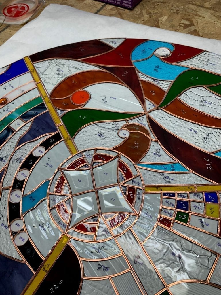 A close-up view of a large circular stained glass panel in progress, laid out on a white surface over a workbench. The panel features an intricate, abstract radial design with a central starburst motif made from clear beveled glass squares. Numerous hand-cut pieces of colored and textured art glass, including vibrant blues, greens, reds, browns, yellows, and clear crackle textures, are visible, many with numerical annotations. All glass pieces are meticulously wrapped with copper foil, ready for soldering.