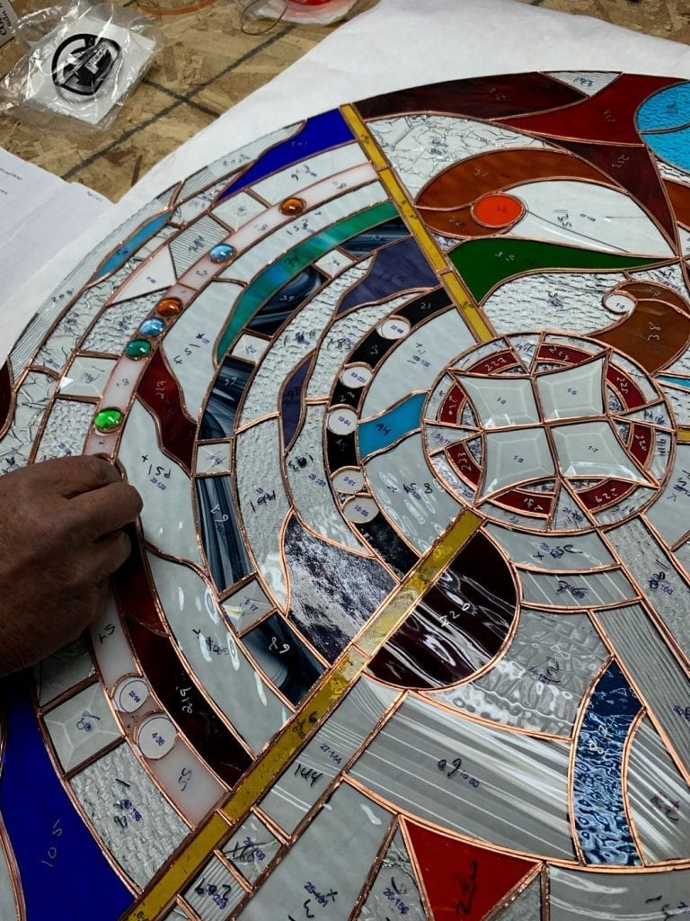 Overhead view of a large, circular stained glass panel under construction, featuring a complex geometric design with clear, white, blue, red, green, and amber glass pieces. An artisan's hand is adjusting one of the pieces, and the panel is meticulously joined by copper foil, with embedded glass jewels visible throughout.