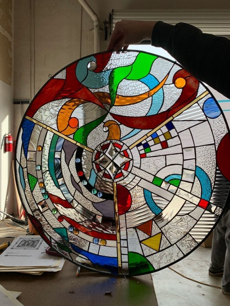 A circular, leaded stained glass panel held up against a window, showcasing a vibrant, abstract, and geometric design. The panel features a central clear beveled starburst radiating outward, dividing the circle into quadrants. It incorporates a rich palette of reds, blues, greens, oranges, and yellows, alongside various clear textured and beveled glass pieces. Small, round colored glass jewels are embedded in one section, while another displays small geometric colored squares. The overall design blends organic, flowing lines with sharp, angular sections, creating a dynamic interplay of color and light.