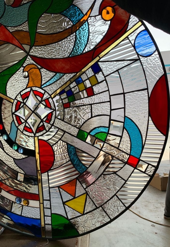 A vibrant, circular leaded stained glass panel featuring a complex abstract and geometric design. It includes a central clear beveled starburst, surrounded by diverse textured and clear glass, along with colorful red, blue, green, yellow, orange, and iridescent elements. Gold-tone lines and small facet-cut jewels add to the dynamic, radiant composition.