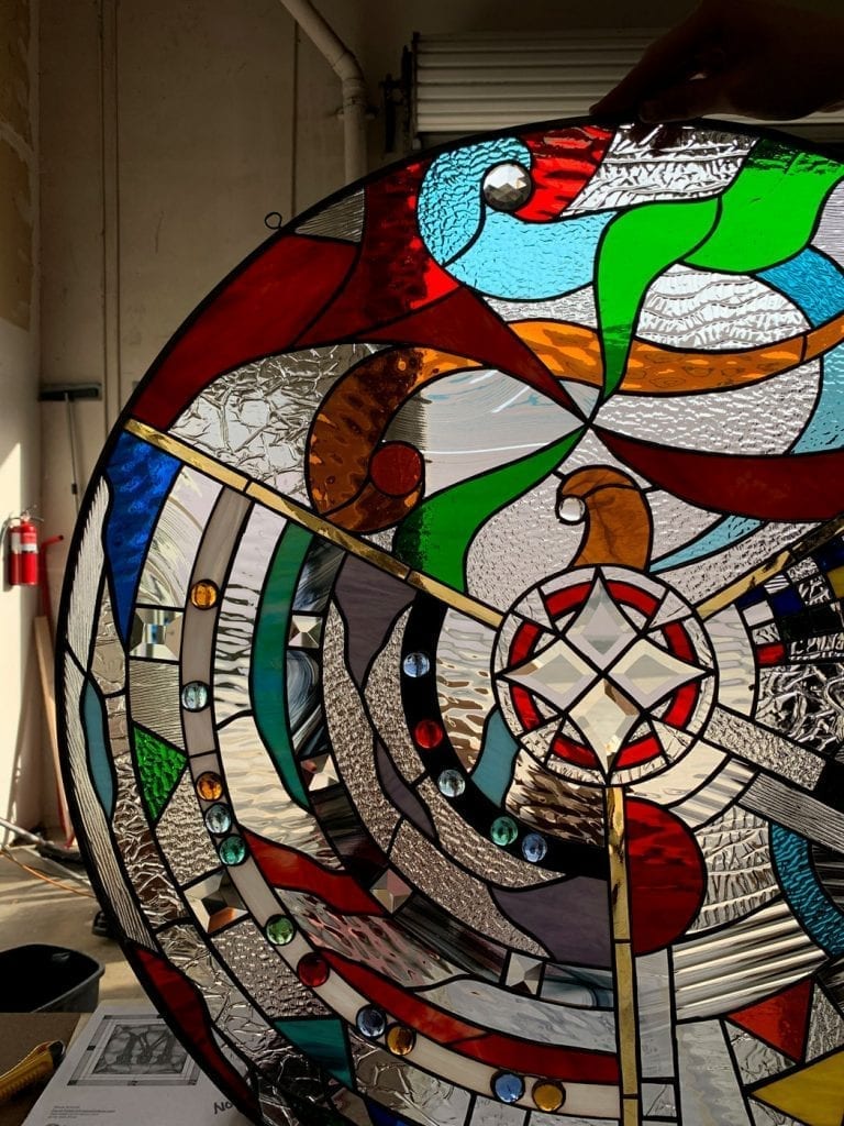 A large, circular stained glass panel features a vibrant, abstract design with swirling patterns in red, blue, green, orange, and clear textured glass. It includes a central geometric medallion with clear bevels, colorful glass jewels, and brass-colored lines, all set in black lead came.