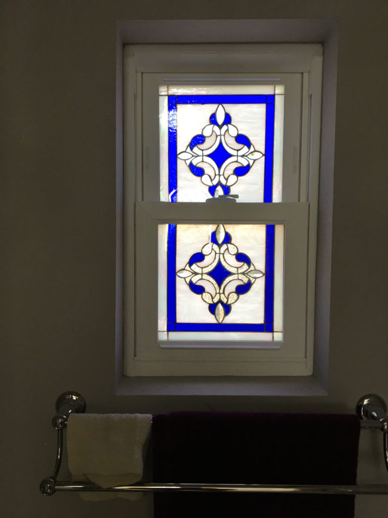 A rectangular double-hung window featuring two identical leaded stained glass panels. Each panel has a central blue quatrefoil design surrounded by clear textured and beveled glass, all within a vibrant blue border. The window is housed in a white frame, with a towel rack below.