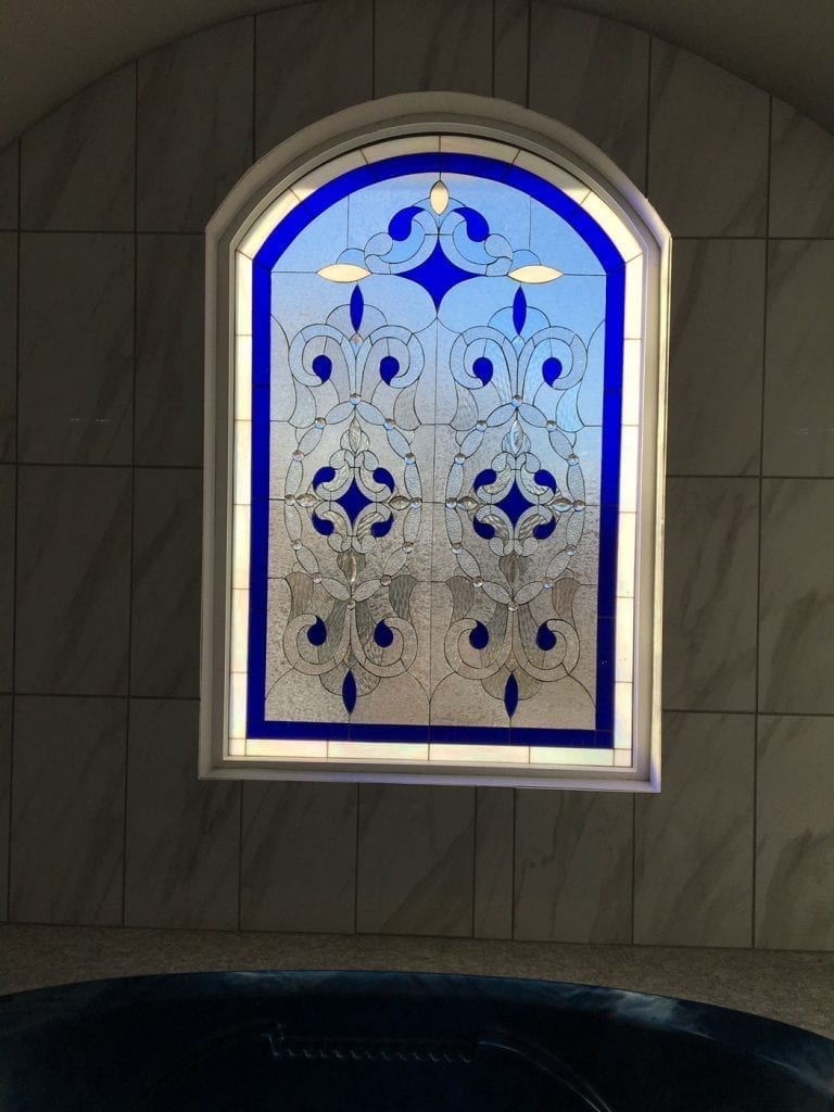 An arched stained glass window features a symmetrical, ornate design with deep royal blue glass accents and clear, beveled, and textured glass panels. The intricate pattern includes scrolls, diamond shapes, and stylized florets, all outlined in lead. The window is set into a light-colored tiled wall above a dark blue tub or jacuzzi, illuminated from behind to highlight its vibrant blue and clear textures.