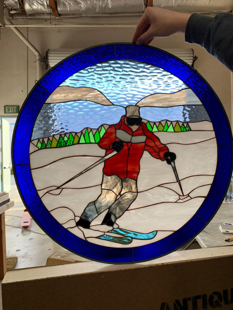 A round leaded stained glass panel depicting a skier in a dynamic pose. The skier, wearing a red jacket, grey pants, and black goggles, glides down a snowy slope with light blue skis. In the background, green triangular trees line the snow, with light brown mountains and textured light blue water and sky above. A vibrant, textured dark blue glass border frames the entire scene.