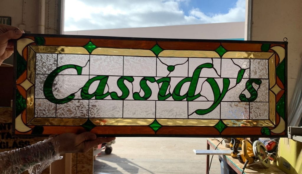 A rectangular leaded stained glass panel displaying the name 'Cassidy's' in vibrant green script. The background is clear textured glass, and the ornate border features geometric patterns in gold, amber, and green diamond-shaped glass.
