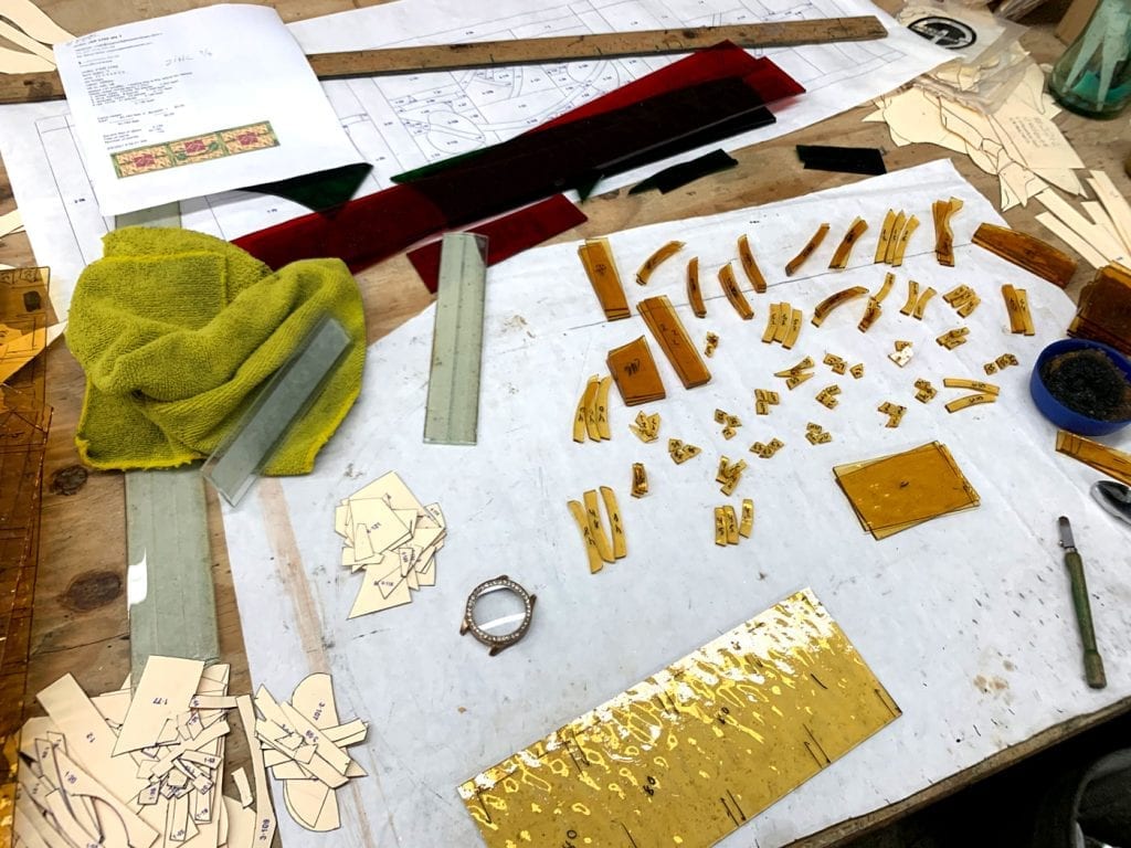 Overhead view of a busy stained glass artist's workbench, showcasing the intricate crafting process. On a white surface, numerous small, individually cut and numbered pieces of amber-colored glass are meticulously laid out, ready for assembly. Surrounding them are detailed paper patterns and templates, larger uncut sheets of vibrant red, dark, and textured amber glass, a precision glass cutting tool, a ruler, and a soft yellow cloth, all illustrating the meticulous steps involved in creating a custom stained glass panel.