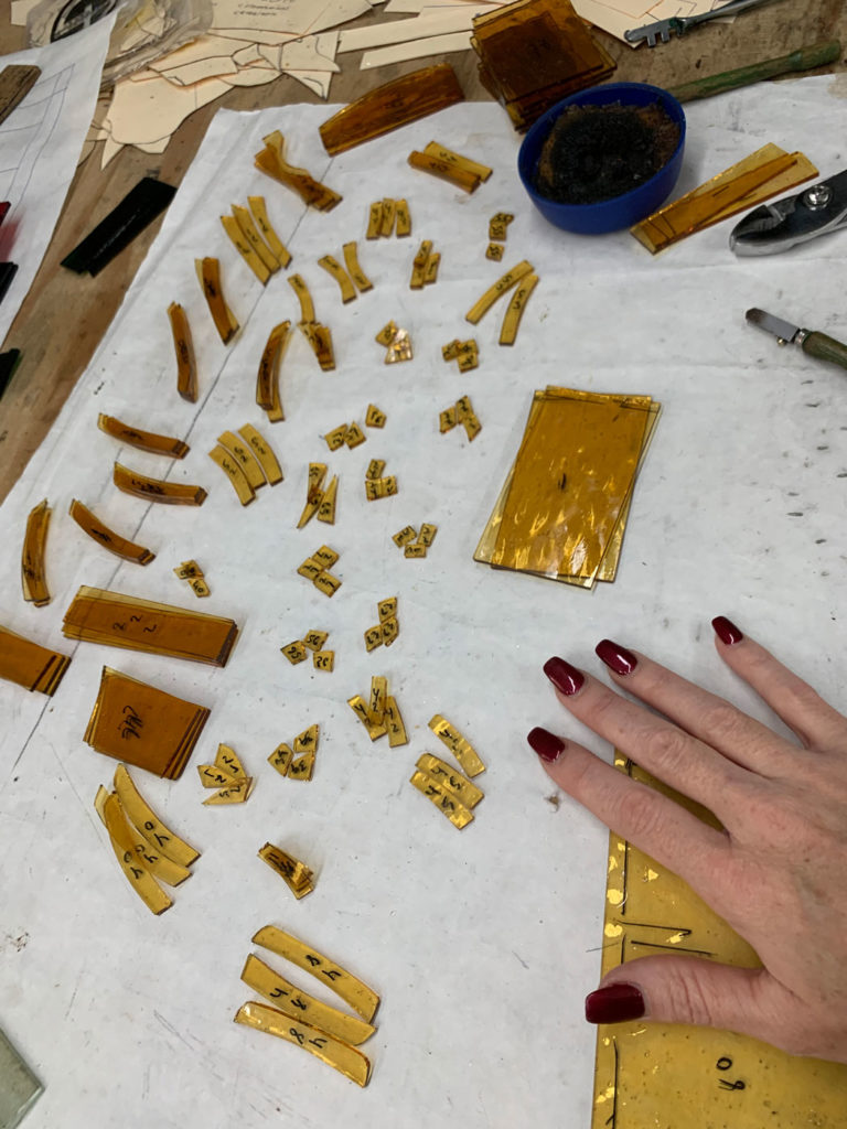 Overhead view of a stained glass artisan's workspace, showing numerous amber-colored glass pieces of various sizes, many marked with black numbers, laid out on a white protective sheet. A hand with dark red nail polish rests on a larger piece of amber glass in the foreground. In the background are paper patterns, glass cutting tools (like a glass cutter and grozing pliers), and a blue bowl containing a dark substance.