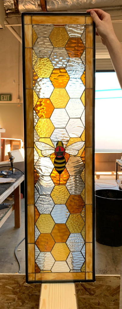 A tall, rectangular stained glass panel featuring a vibrant honeycomb pattern in various shades of amber, golden yellow, and clear textured glass. A detailed bumblebee with yellow and black stripes, a red head, and translucent wings is prominently centered within the hexagonal pattern. The panel has a light brown glass border and a dark metal frame.