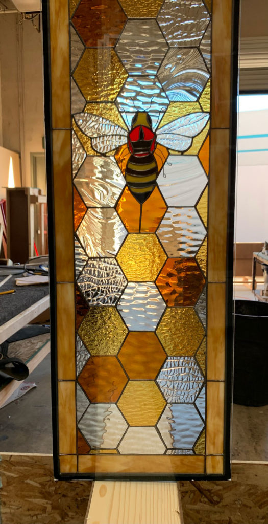 A rectangular stained glass panel featuring a prominent black and yellow bumblebee with red accents at its center. The bee is surrounded by a leaded honeycomb pattern, with hexagonal cells filled with various clear, amber, and golden textured glass. The entire panel is framed by a solid light amber border.