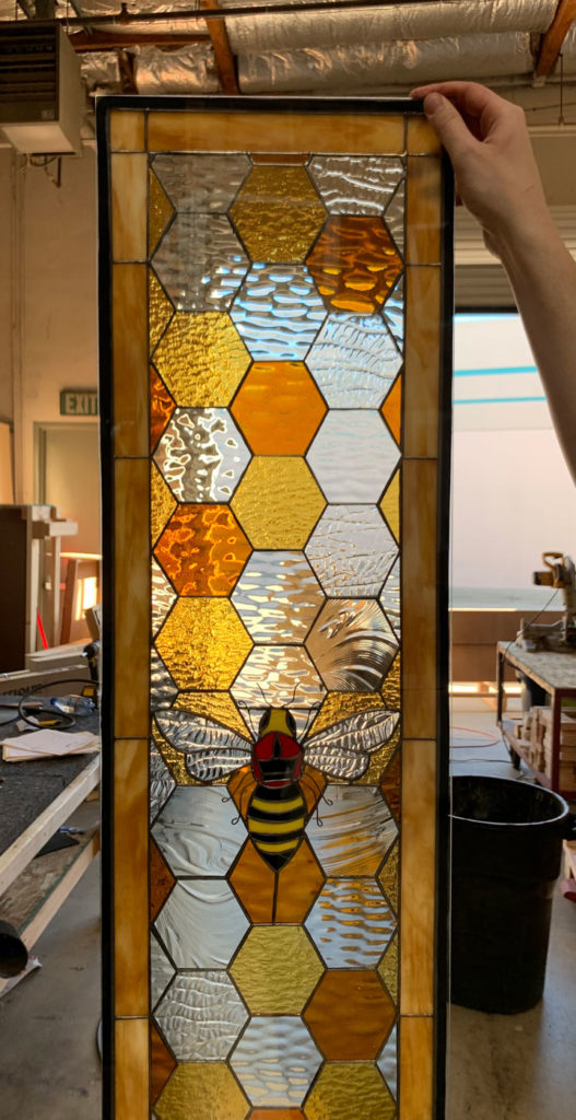 A vertical rectangular leaded stained glass panel depicting a vibrant, striped bee with red accents on its head, resting on an intricate honeycomb design. The honeycomb features a mix of textured clear, golden yellow, and amber hexagonal glass pieces, framed by a rich amber border.