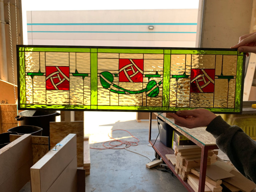 A rectangular leaded stained glass panel held by hands, featuring a vibrant lime green border. The main field is textured golden-yellow glass, accented by three red square sections containing intricate white abstract swirling patterns, interconnected by a flowing green ribbon design, all back-lit.