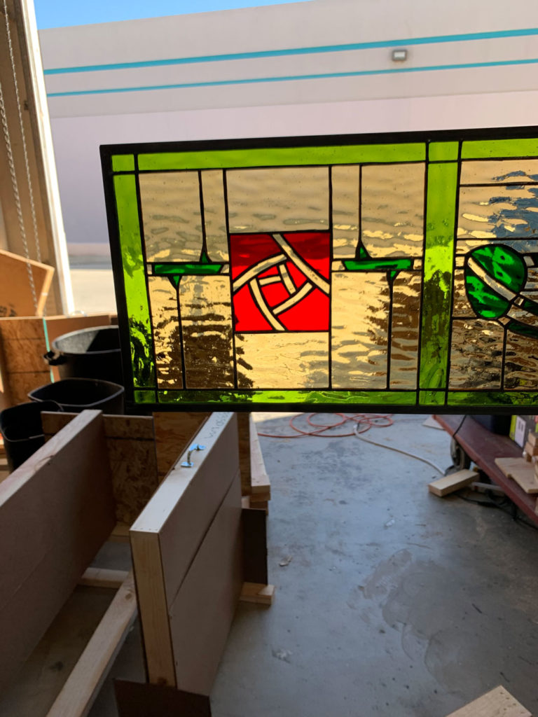 A rectangular stained glass panel featuring a bright green border, clear textured glass, and a central red square with an intricate white geometric knot design. Additional green glass elements are on the right.