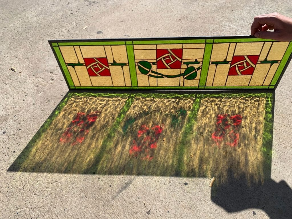 A rectangular Prairie-style leaded stained glass panel featuring a repeating pattern of three stylized red roses enclosed in geometric cream squares, interconnected by vivid green lineal accents and organic leaves. The panel, held vertically, casts intricate, colored shadows of its design onto a light gray concrete surface under bright sunlight, showcasing its vibrant red, green, and cream glass.