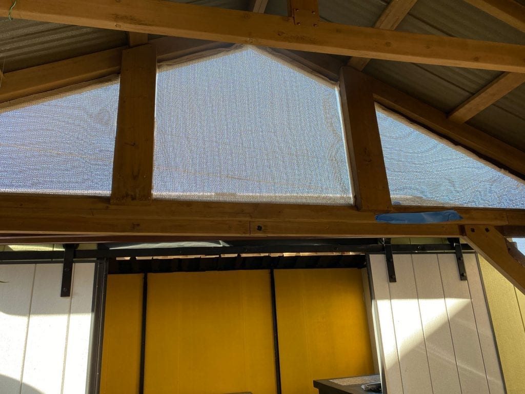 A gabled wooden roof structure featuring mesh screens in the triangular and trapezoidal window openings, ideal for custom stained glass panels. Below, a yellow interior wall is visible next to a white sliding barn door, creating a rustic outdoor living area.