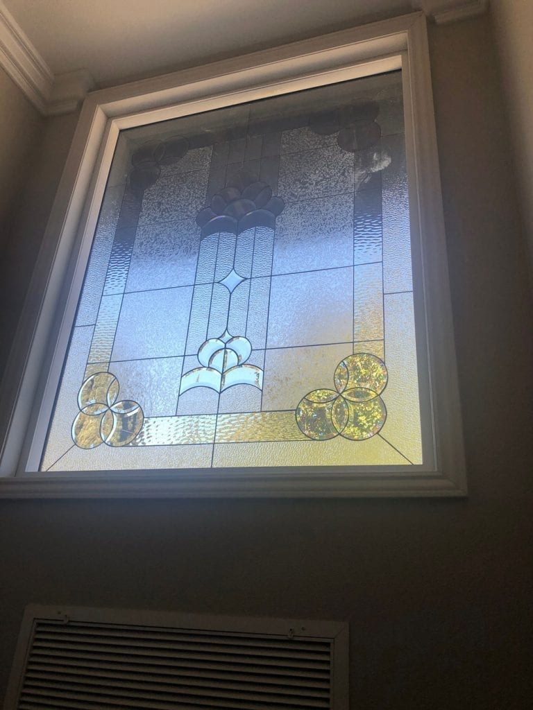 A rectangular leaded stained glass window panel, set in a white frame, featuring a central Art Nouveau-inspired design. The panel is primarily composed of clear textured glass, showcasing a prominent stylized floral or urn motif at the center, flanked by a star-like element. The bottom corners each display a cluster of three overlapping circles made from clear and warm amber-toned textured glass. Subtle cloud-like patterns appear in the upper corners, and the glass diffuses natural light with hints of blue and gray.