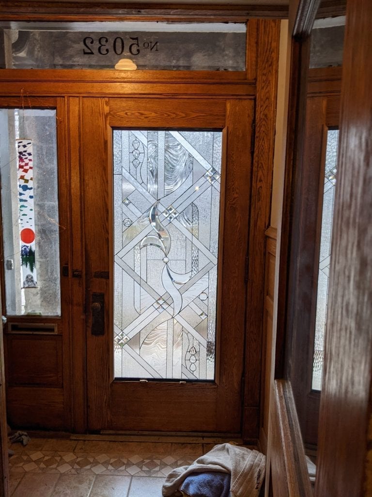 An interior view of a dark wood entry door featuring a large, clear stained glass panel. The leaded glass showcases an intricate Art Deco geometric pattern composed of various textured and beveled clear glass pieces, including a prominent central organic swirl. Above the door, a transom window displays the reversed house numbers 'Nº 2035'. A matching wood sidelight is visible to the left.