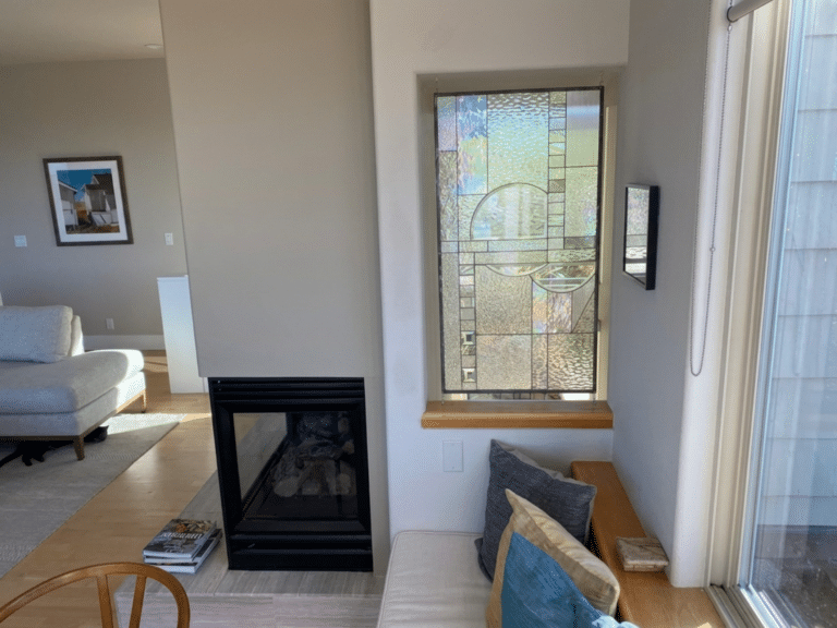 A rectangular, leaded stained glass panel featuring an abstract geometric design with various clear textured and iridescent glass pieces, and two circular elements, installed in a wall opening between a fireplace and a large window.