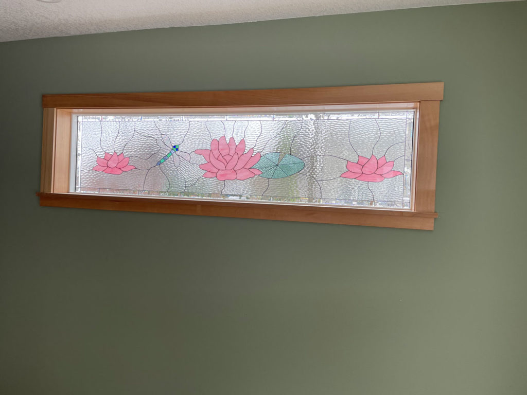 A horizontal stained glass transom panel, set in a natural light wood frame, featuring a vibrant pond scene with three pink lotus flowers, a green lily pad, and a delicate blue and green dragonfly, all set against a textured clear glass background.