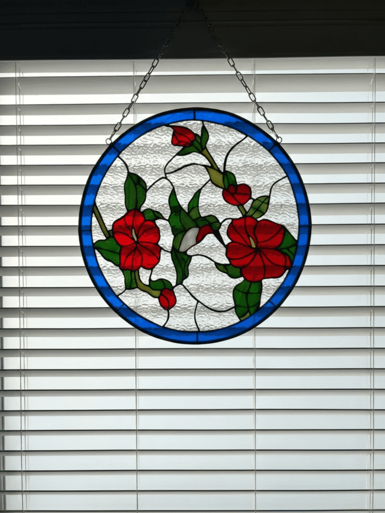 A circular stained glass panel depicting a white hummingbird feeding among red flowers and green leaves, set against a textured clear glass background with a blue outer border. The panel is hanging by chains in front of white horizontal window blinds.