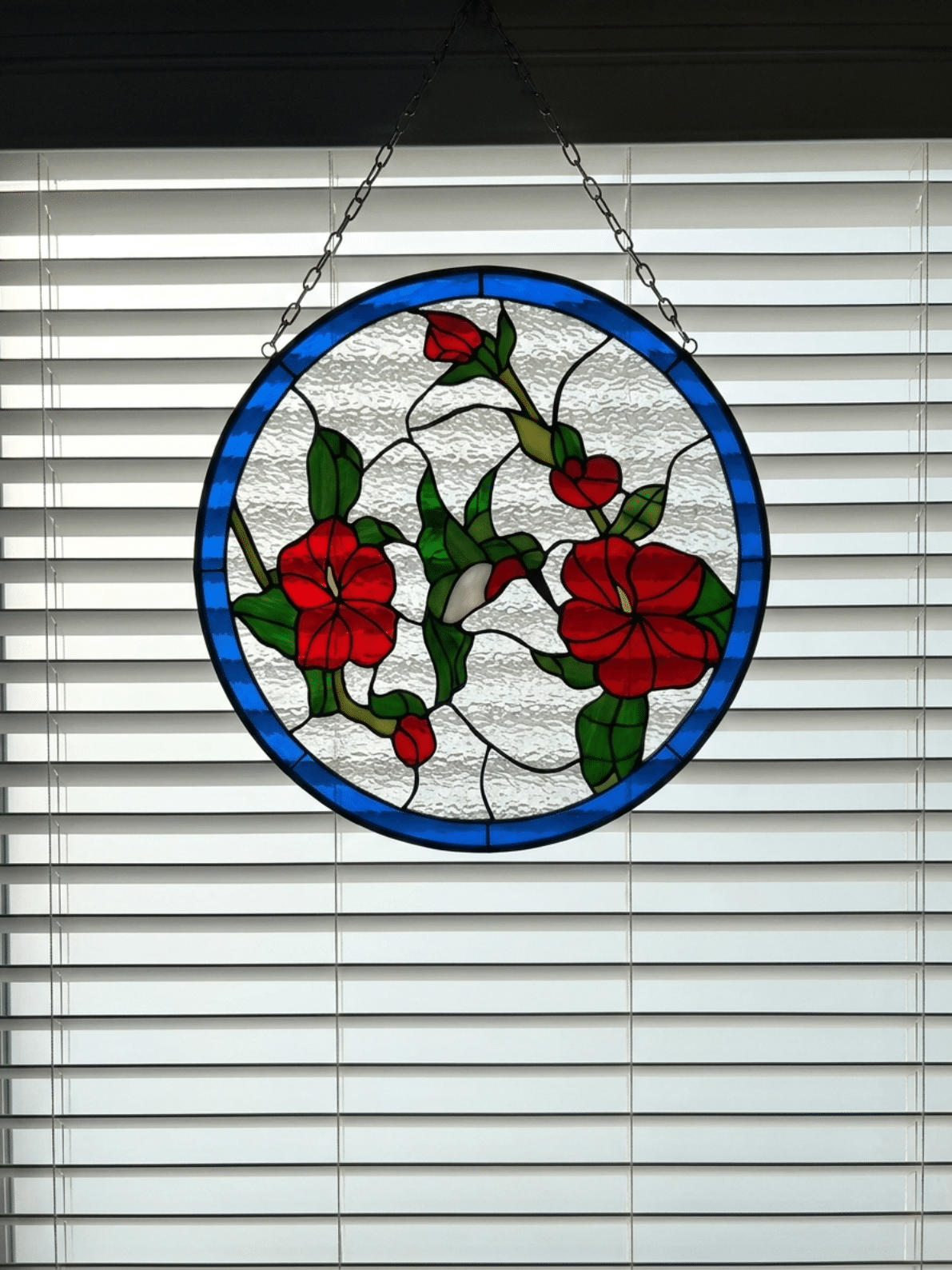 A circular stained glass panel depicting a white hummingbird feeding among red flowers and green leaves, set against a textured clear glass background with a blue outer border. The panel is hanging by chains in front of white horizontal window blinds.