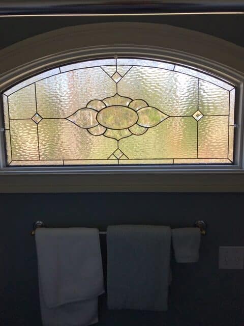 An arched leaded stained glass window featuring a central geometric floral medallion design with clear beveled and textured glass, providing privacy in a bathroom setting.