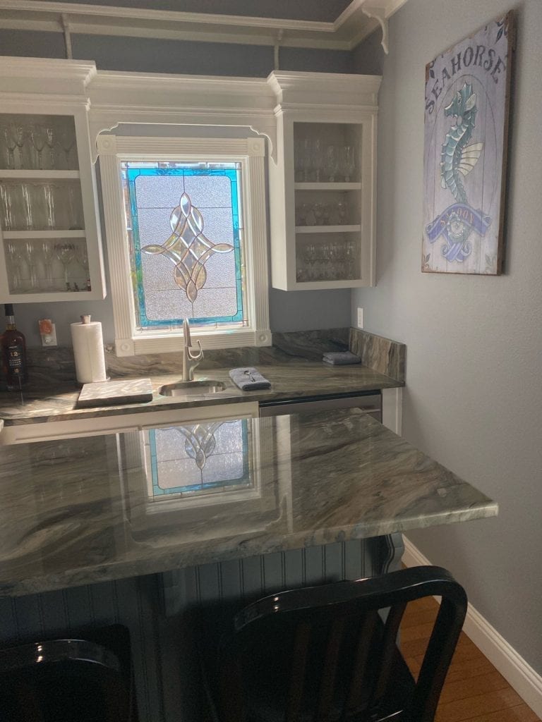 A rectangular stained glass window panel with a central clear and amber geometric design, bordered by vibrant blue glass, installed above a kitchen sink with ornate white trim. White upper cabinets flank the window, and a reflective granite countertop is visible below.