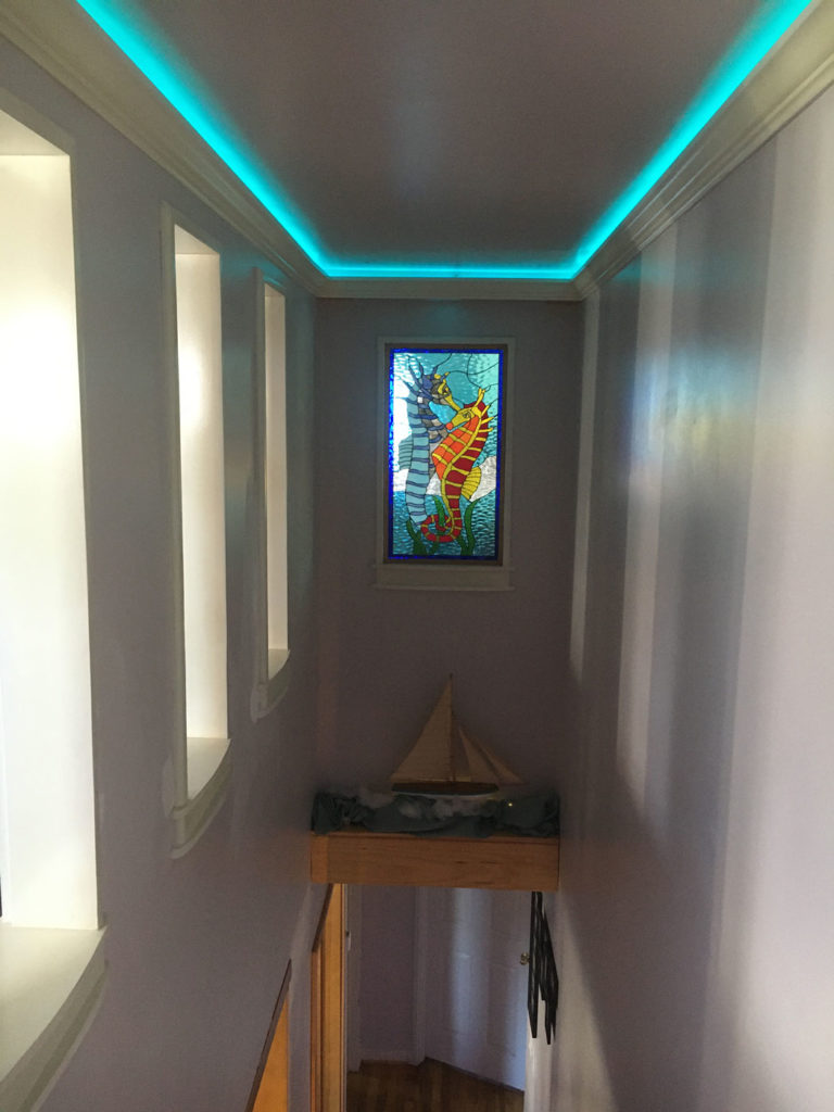 A rectangular, vertically oriented leaded stained glass panel depicting a large, detailed orange and yellow seahorse swimming amongst blue wavy water and green marine plants. The panel is installed in a light-colored wall, illuminated by a glowing turquoise LED strip light recessed in the ceiling molding, creating an ambient oceanic atmosphere. Below the panel, a wooden model sailboat rests on a small shelf.