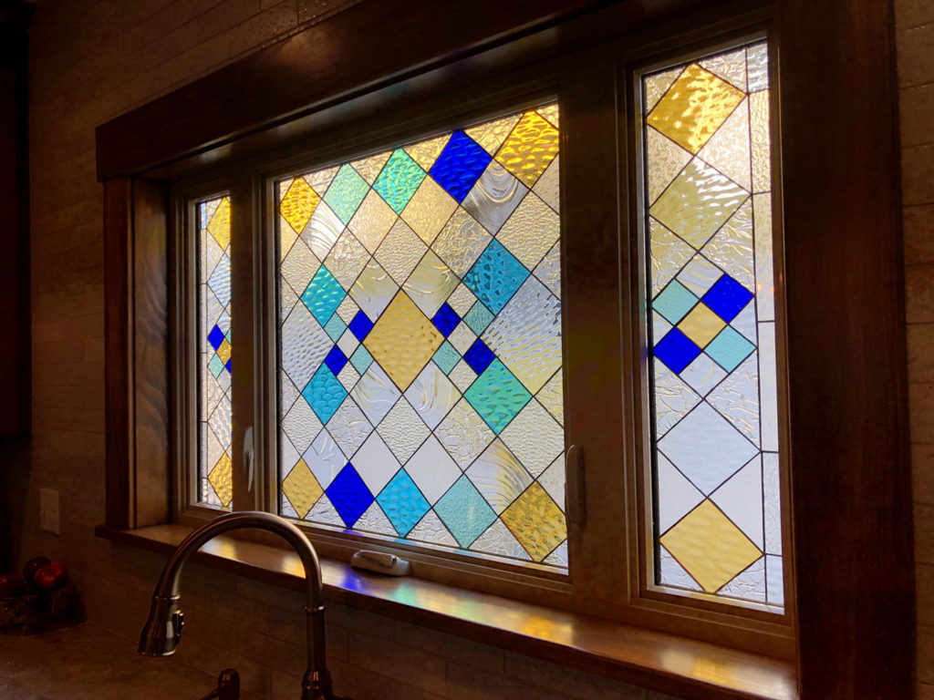 A multi-panel leaded stained glass window installed above a kitchen sink, featuring a geometric pattern of diamond shapes in clear textured, yellow, light blue, and deep blue glass. The sunlight illuminates the textured glass, casting colorful reflections.