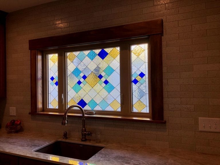 A rectangular stained glass panel installed in a kitchen window above a sink, featuring a vibrant geometric diamond pattern crafted from clear textured glass, soft yellow, light blue, and brilliant blue diamond shapes.