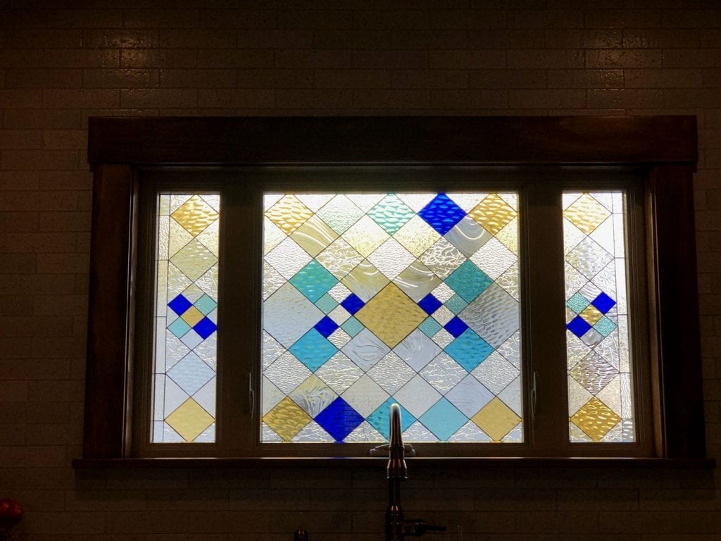A rectangular stained glass window, set in a dark wood frame, featuring a modern geometric design composed entirely of diamond shapes. The panel showcases clear textured glass interspersed with light blue, golden yellow, and royal blue diamond pieces, arranged in a repeating diagonal grid. The main central section is wider, flanked by two narrower side panels, all sharing the same diamond pattern.