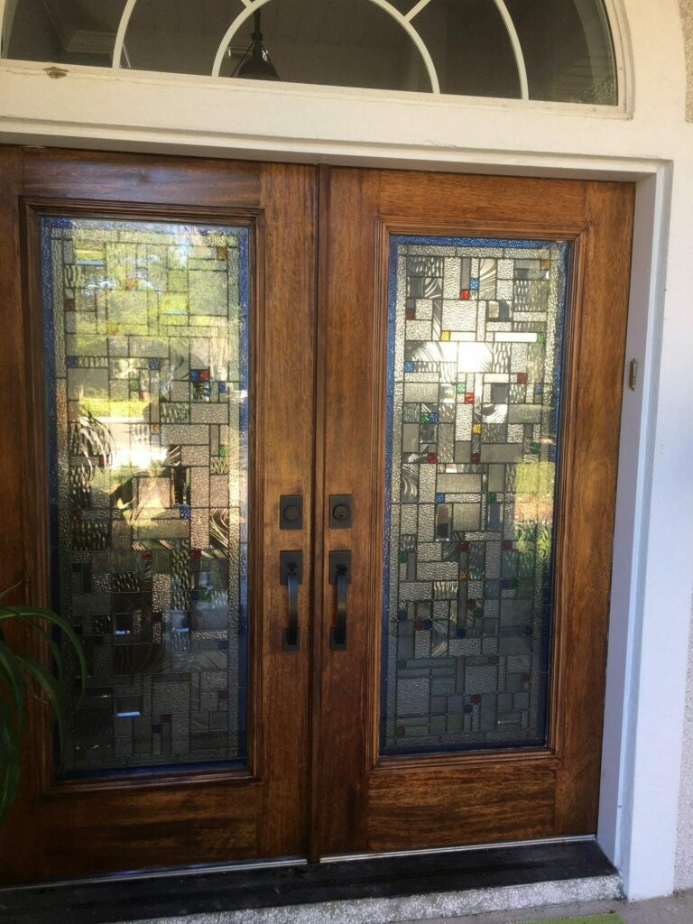 A pair of warm-toned wooden double entry doors, each fitted with a full-length stained glass panel. The panels feature a contemporary geometric design of clear and various textured glass, accented with small, colorful square pieces in red, blue, green, and yellow. An arched transom window is visible above the doors.