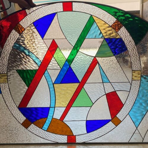 An arched leaded stained glass panel featuring a vibrant abstract geometric design. It has a central circular pattern composed of red, blue, green, and yellow triangles and lines, surrounded by a diverse array of clear textured and reeded glass, with additional colorful and clear segments in the arch's outer areas.