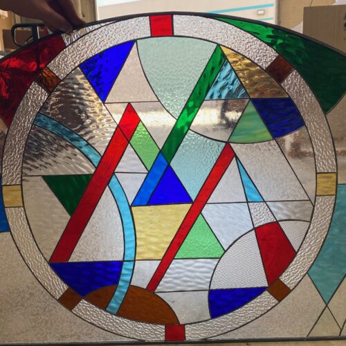 An arched stained glass panel featuring a large central circular motif filled with an abstract geometric design of interconnected triangles and curved segments in vibrant red, blue, green, yellow, and amber, interspersed with various clear textured glasses like hammered, ripple, and streaky patterns, all joined by dark leading. The outer arched sections also contain clear textured glass and solid color segments.