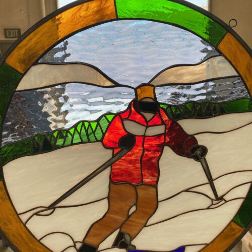 A round leaded stained glass panel depicting a vibrant scene of a skier in a red jacket and brown pants on a snow-covered mountain. The background features a blue and white sky, grey mountains, and green evergreen trees. The circular border is segmented with rich green and amber glass.