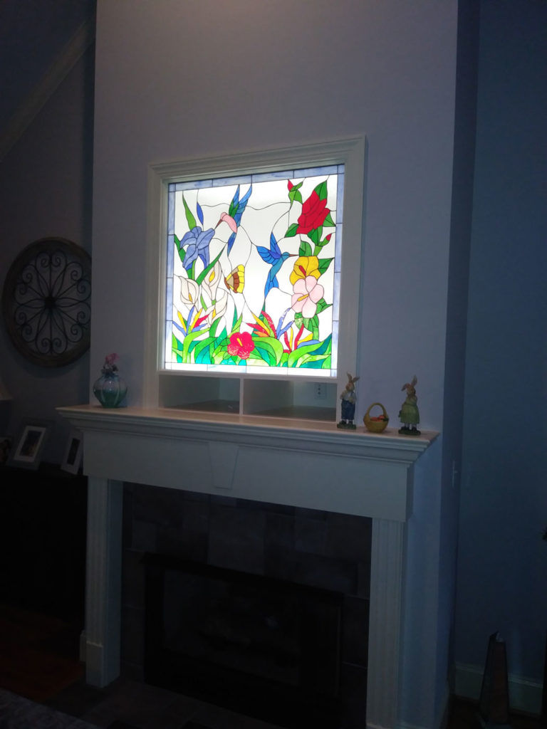 A rectangular, brightly backlit stained glass window installed above a white fireplace mantel. The vibrant design features two hummingbirds – one blue, one pink – amidst a colorful array of tropical flowers, including large red blossoms, purple irises, yellow blooms, and white calla lilies, all set against a clear background. The window is framed in white trim.
