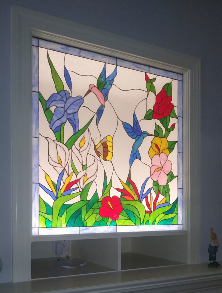 A rectangular stained glass panel depicting a lively tropical garden scene. It features two hummingbirds, one butterfly, and an assortment of colorful flowers including blue irises, white calla lilies, red and pink hibiscus, yellow flowers, and orange bird of paradise, all surrounded by green foliage against a clear textured glass background, finished with a blue border.