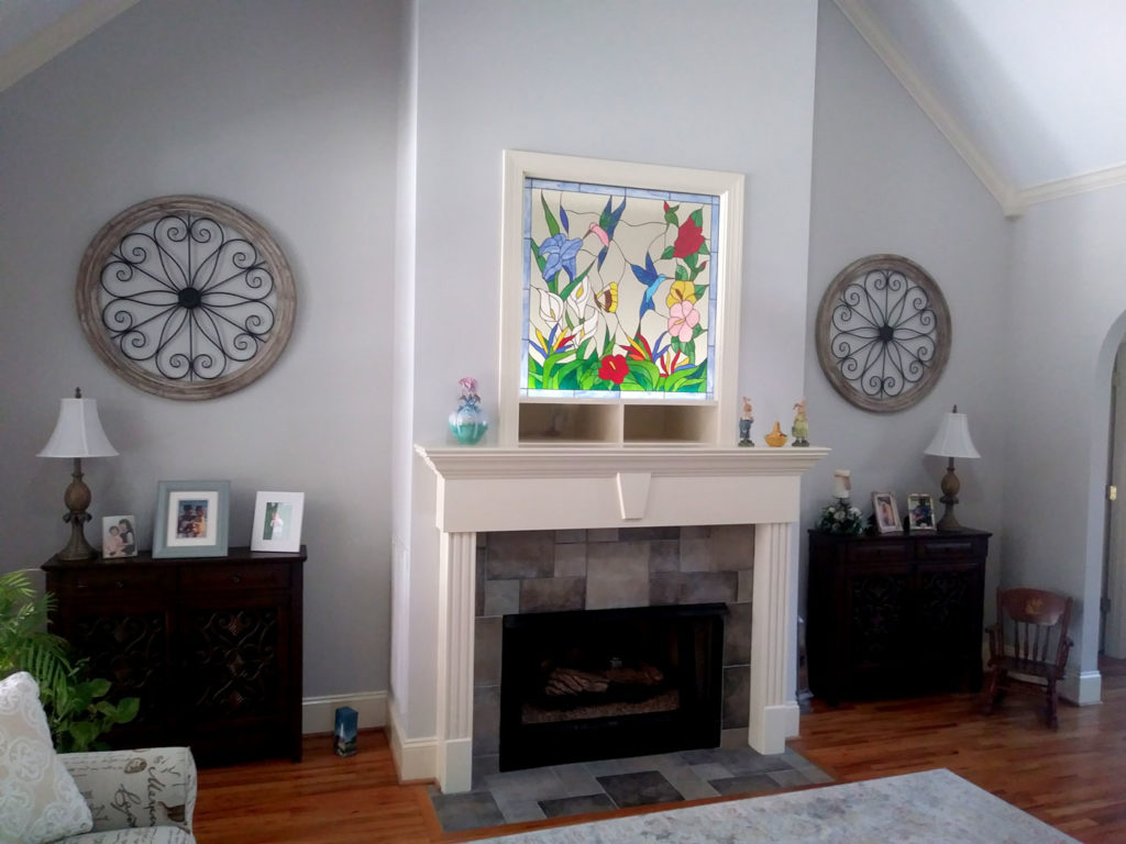 A rectangular stained glass panel showcasing a lively nature scene with multiple colorful hummingbirds, butterflies, and an assortment of flowers in red, yellow, blue, and pink, accented by green leaves. The panel is set in a white frame, mounted above a cream-colored fireplace mantel.
