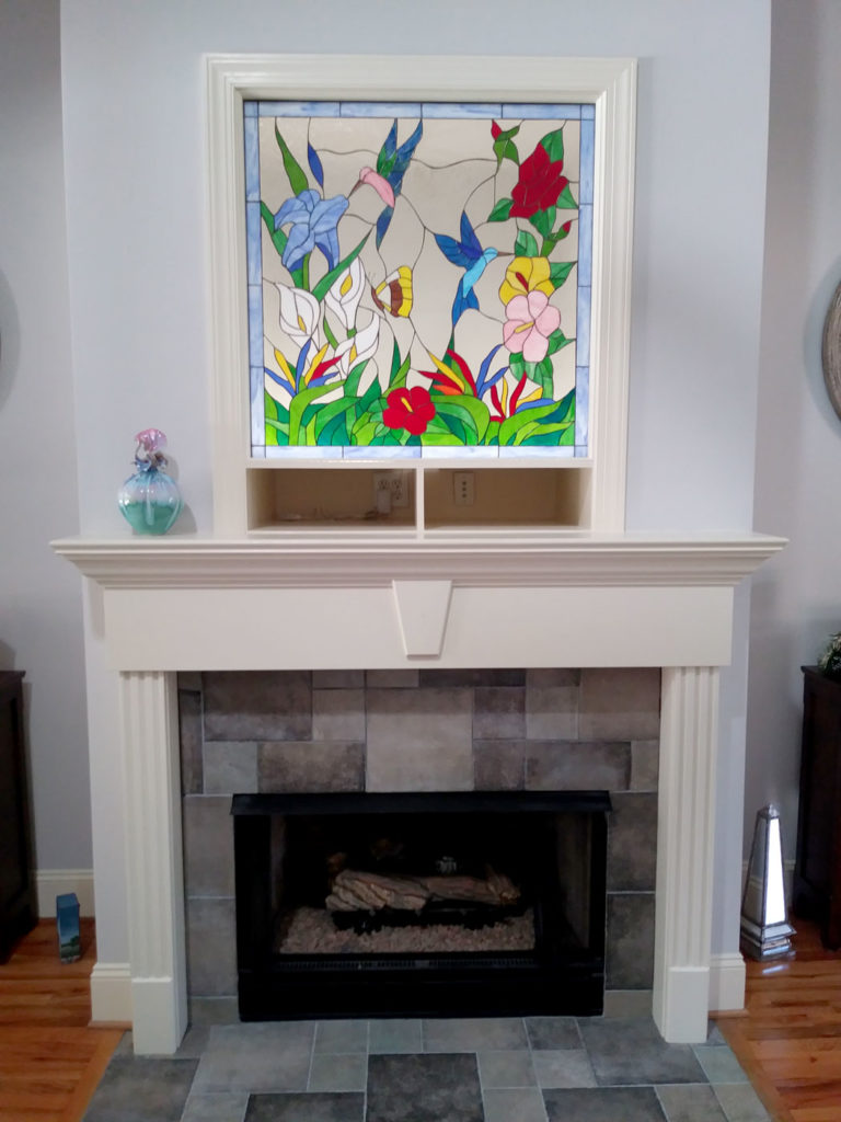 A rectangular stained glass panel showcasing a vibrant tropical garden scene with two hummingbirds, a yellow butterfly, and various colorful flowers including red hibiscus, blue irises, white calla lilies, and yellow blossoms, all set against a clear textured background, installed above a white fireplace mantel.