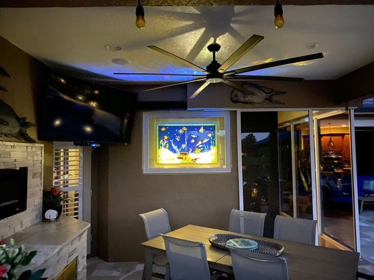 A brightly back-lit, rectangular stained glass panel showcasing a vibrant underwater scene with yellow, blue, and red tropical fish swimming amidst green and red coral reefs and blue-green seaweed, set in a white frame on a dark wall in an outdoor dining area.