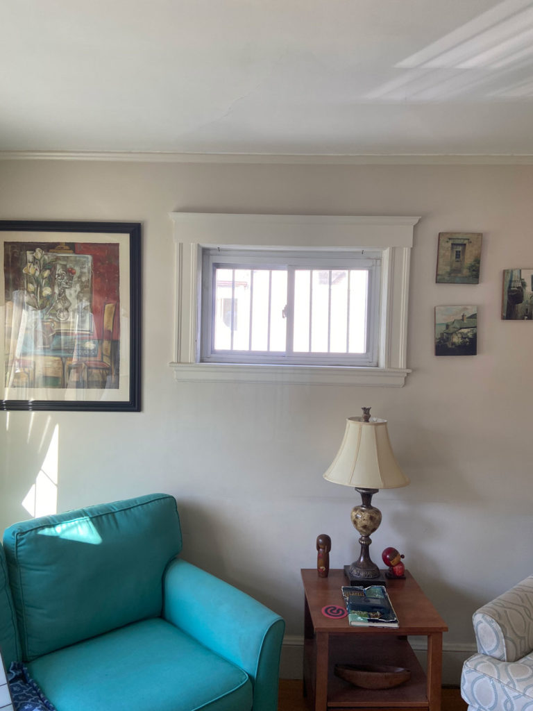 A rectangular leaded stained glass panel, featuring clear textured glass and elegant beveled accents, installed within a white-trimmed window. The window is positioned above a wooden side table with a lamp, and next to a vibrant turquoise armchair, in a sunlit living room.