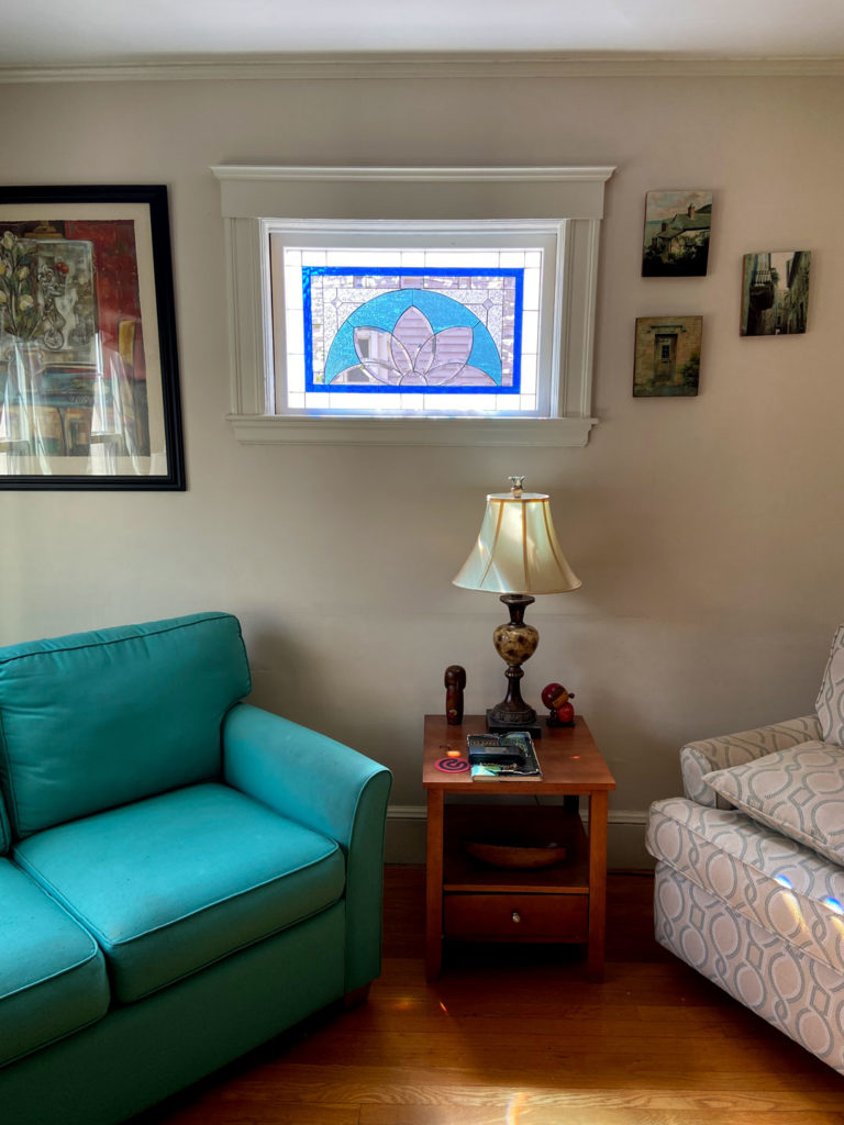 A rectangular leaded stained glass panel with a vibrant blue outer border, featuring a central semi-circular design of clear and textured glass segments forming a stylized lotus or floral pattern. The panel is installed within a white window frame in a sunlit living room.