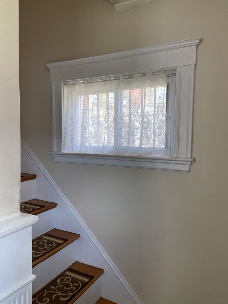 A rectangular window with decorative white lace curtains and prominent white trim, positioned above a wooden staircase featuring brown patterned runners, against a neutral-colored wall. This space is perfectly suited for a custom stained glass installation.