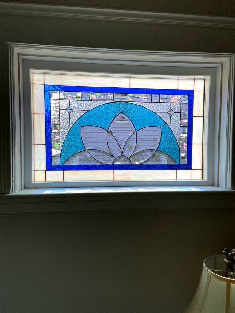 A rectangular leaded stained glass window panel, installed in a white frame. It features a central light blue floral or lotus-like motif made of textured glass, surrounded by a wide royal blue border, and an outer border of clear iridescent rectangular glass pieces. The background through the clear areas shows an outdoor residential scene.