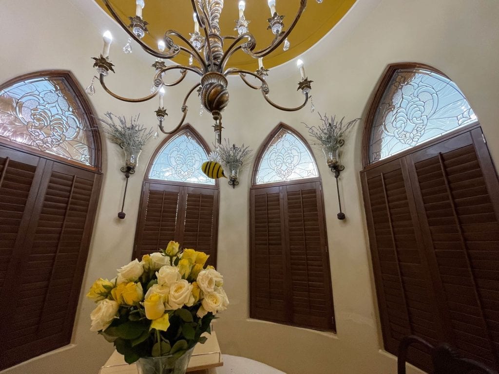 A set of four arched windows, each featuring a clear, textured, and beveled leaded stained glass panel with an intricate scrollwork design in the upper portion, positioned above dark brown louvered plantation shutters. A chandelier hangs from the ceiling, and yellow and white roses are on a table in the foreground.