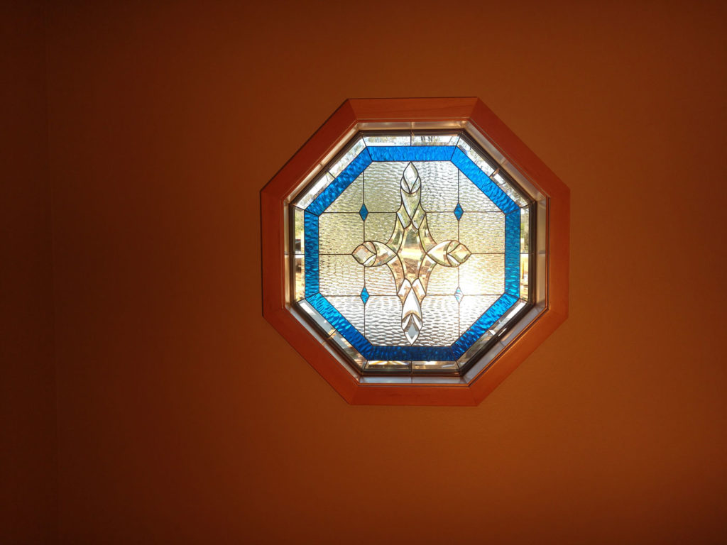 An octagonal stained glass window with a light wood frame. The clear, textured, and beveled glass panel features a central stylized cross or fleur-de-lis design, bordered by vibrant blue textured glass.