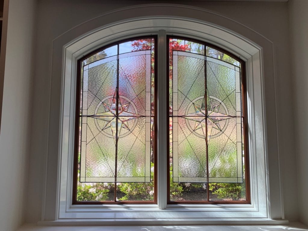 A large arched window composed of two clear stained glass panels. Each panel displays a central compass rose design with clear beveled glass, framed by various clear textured glass patterns and diagonal lead lines. The dark window frames are set within a white arched architectural casing, with green and red foliage visible outside.